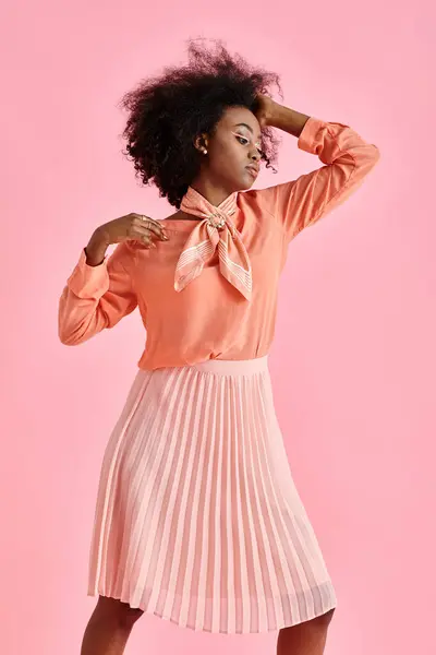 African american woman in peach blouse, midi skirt and scarf adjusting curly hair on pink backdrop — Stock Photo
