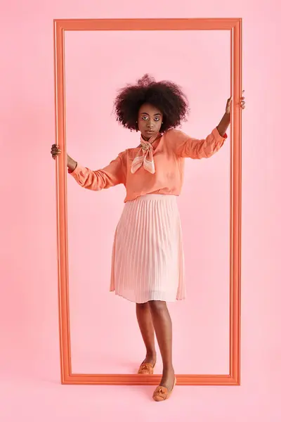 Молода афроамериканка в персиковій блузці і спідниці міді страйкує позу біля рамки на рожевому тлі — стокове фото