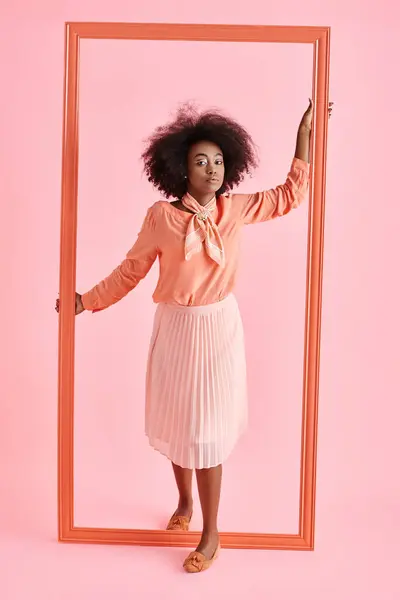 Pretty african american woman in peach blouse and midi skirt strikes pose near frame on pastel pink — Stock Photo