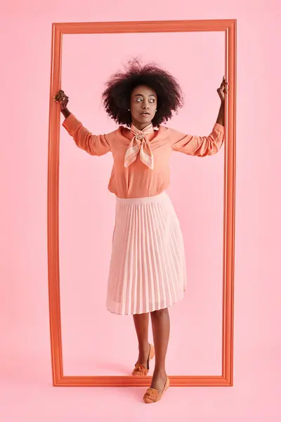 Молода афроамериканка в персиковій блузці і спідниці міді страйкує позу біля рамки на пастельному рожевому — стокове фото