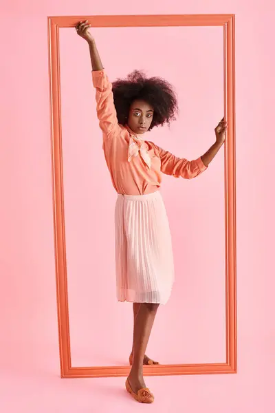 Joven afroamericana mujer en blusa de melocotón y falda midi posando cerca de marco sobre fondo rosa - foto de stock
