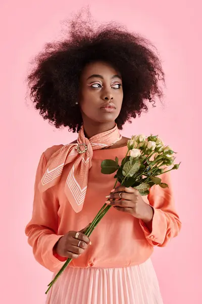 Chica afroamericana soñadora en blusa de pelusa de melocotón sosteniendo flores y mirando hacia otro lado en el telón de fondo rosa - foto de stock