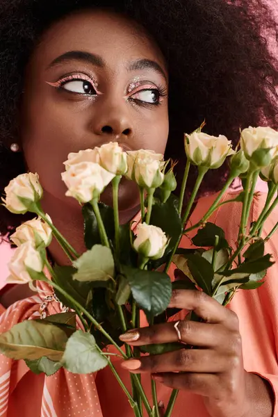 Mujer afroamericana en traje de pelusa de melocotón sosteniendo flores y mirando hacia otro lado en el telón de fondo rosa - foto de stock