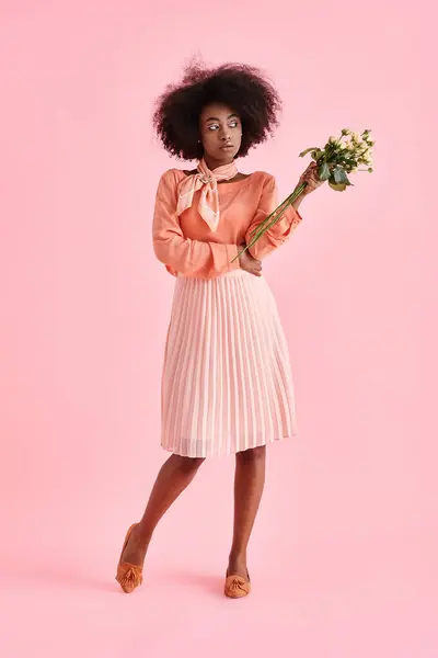 Mulher americana africana em pêssego fuzz roupa segurando flores e olhando para o fundo rosa — Fotografia de Stock