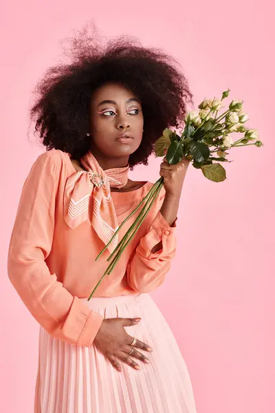 Mujer afroamericana en traje de pelusa de melocotón sosteniendo flores y mirando hacia otro lado sobre fondo rosa - foto de stock