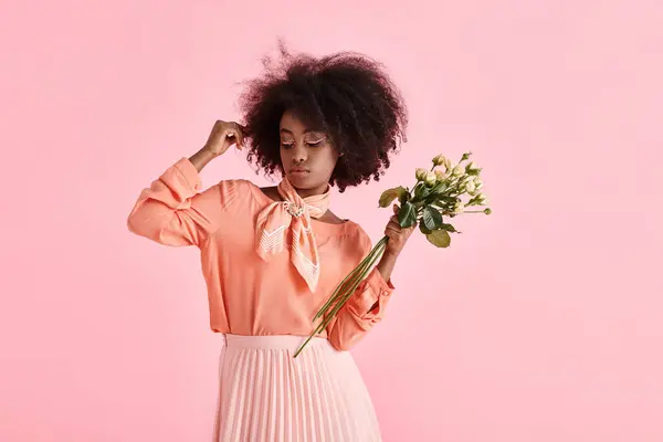 Mujer afroamericana en traje de pelusa de melocotón sosteniendo flores y mirando hacia abajo sobre fondo rosa - foto de stock