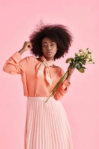 Mujer afroamericana en traje de pelusa de melocotón sosteniendo flores y mirando a la cámara sobre fondo rosa - foto de stock