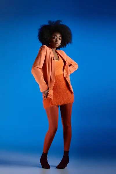 Modelo americano africano bonito en vestido texturizado y chaqueta posando con la mano en la cadera en tacones altos - foto de stock