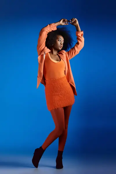 Curly young african american model in textured dress and blazer posing with raised hand on blue — Stock Photo
