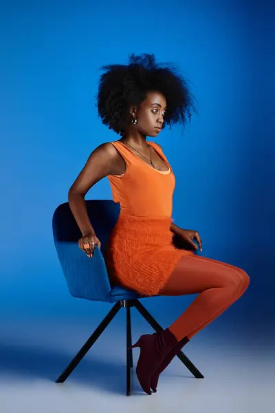Graciosa mulher americana africana em vestido texturizado e salto alto sentado na cadeira no fundo azul — Fotografia de Stock