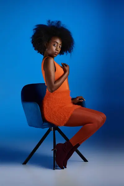 Изящная африканская американская модель в текстурированном платье и на высоких каблуках, сидящая на стуле на синем фоне — стоковое фото