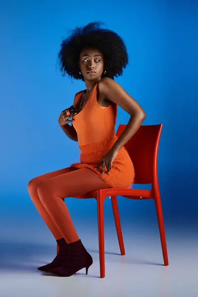 Graceful african american woman in orange dress and high heels sitting on chair on blue backdrop — Stock Photo