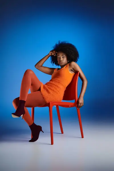 Стильна афроамериканська жінка в помаранчевій сукні і високих підборах сидить на стільці на синьому фоні — стокове фото