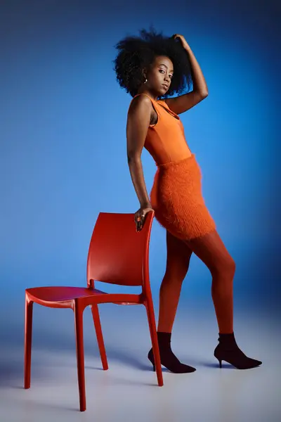 Stylish african american woman in orange dress and high heels posing next to chair on blue backdrop — Stock Photo