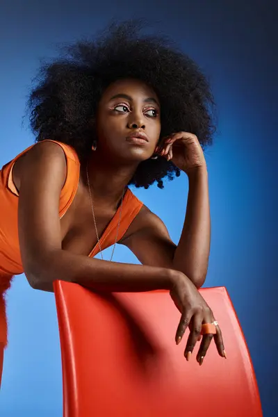 Soñadora mujer afroamericana en vestido de pelusa de melocotón posando al lado de la silla sobre fondo azul vivo - foto de stock