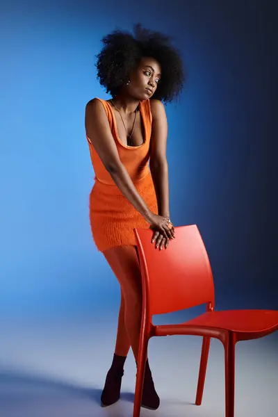 Mujer afroamericana bonita en vestido naranja y tacones altos posando al lado de la silla sobre fondo azul - foto de stock