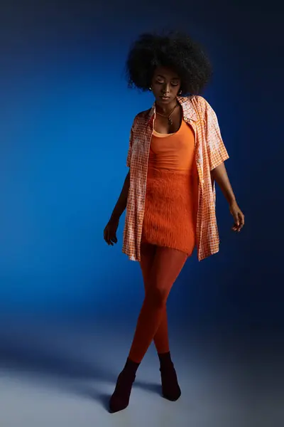 Шикарний вигляд молодої афроамериканки у візерунковій сорочці та помаранчевій сукні на синьому фоні — стокове фото