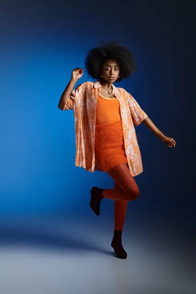 Chic look of african american woman in patterned shirt and orange dress posing on blue background — Stock Photo
