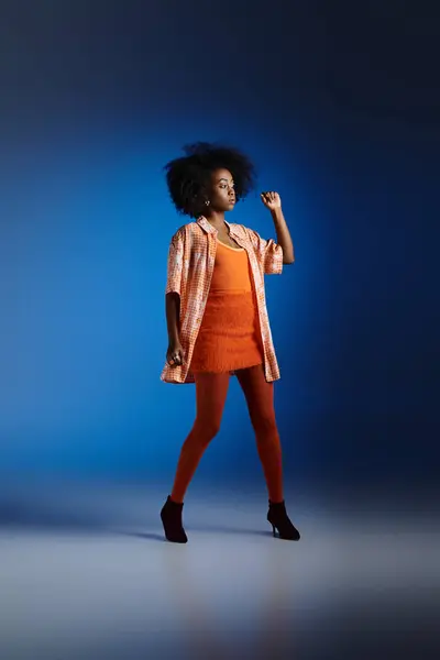 Elegante look de mujer afroamericana en camisa estampada y vestido naranja posando sobre fondo azul - foto de stock
