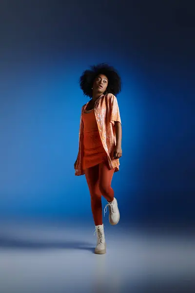 Модний вигляд афроамериканської моделі у візерунковій сорочці та помаранчевій сукні, що позує на синьому фоні — стокове фото