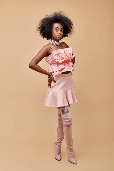 Lockige junge afrikanisch-amerikanische Frau in pfirsichfarbenem Rüschenoberteil und Overknee-Stiefeln auf beigem Hintergrund — Stockfoto