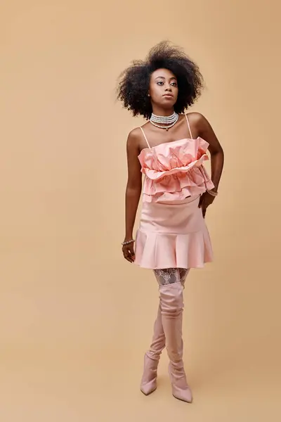 Morena joven afroamericana mujer en melocotón con volantes superior y botas de rodilla sobre fondo beige - foto de stock