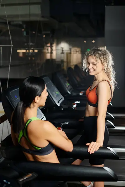 Entrenador femenino atractivo positivo y su cliente en ropa deportiva haciendo ejercicio activamente en la cinta de correr en el gimnasio - foto de stock