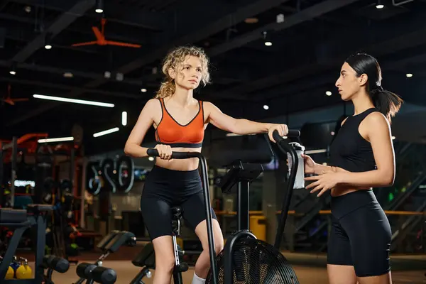 Belle femme énergique utilisant un cross trainer moderne dans la salle de gym à côté de son entraîneur féminin athlétique — Photo de stock