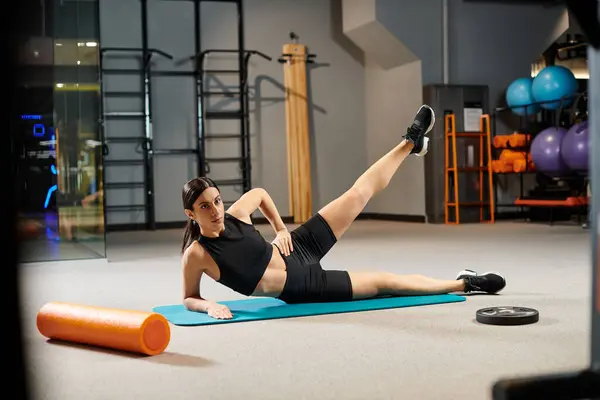 Séduisante femme aux cheveux foncés en tenue de sport noire qui s'étire activement sur un tapis de fitness en salle de sport — Photo de stock