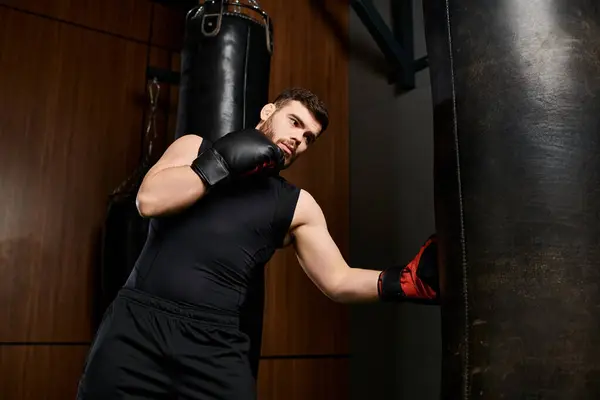 Un homme barbu portant une chemise noire donne des coups de poing à un sac de boxe dans une salle de gym alors qu'il porte des gants de boxe rouges. — Photo de stock