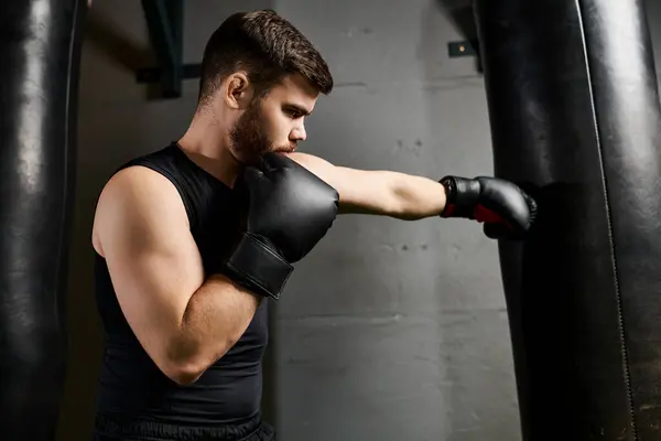 Hombre barbudo guapo con camiseta negra y guantes de boxeo golpea ferozmente una bolsa en un gimnasio. - foto de stock
