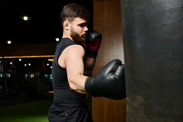 A stylish man with a beard, wearing a black tank top and boxing gloves, is seen punching a bag in a gym. — Stock Photo