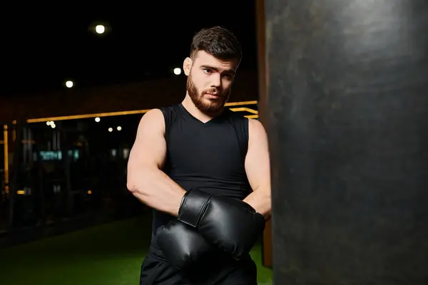 A handsome man with a beard wearing black boxing gloves and a tank top, fiercely punching a bag in a gym. — Stock Photo