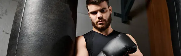 Un guapo barbudo con guantes de boxeo está junto a un saco de boxeo en un gimnasio, listo para una sesión de entrenamiento. - foto de stock