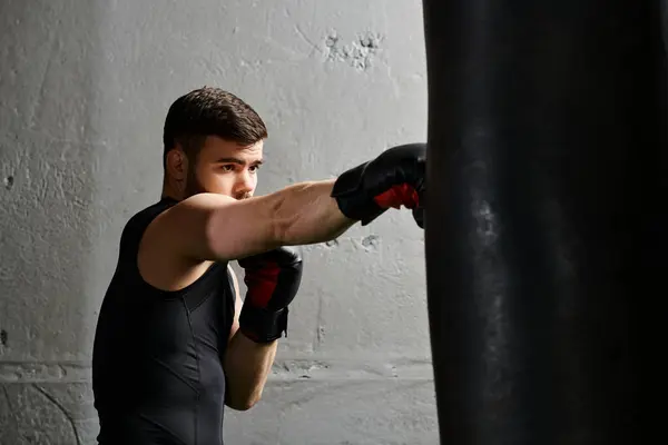 Un hombre guapo con barba con camisa negra y guantes de boxeo negros, golpeando una bolsa en un gimnasio. - foto de stock