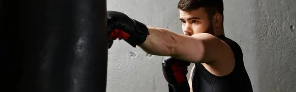 Un hombre guapo con barba que boxea ferozmente en un gimnasio con un saco de boxeo rojo con una camisa negra. - foto de stock