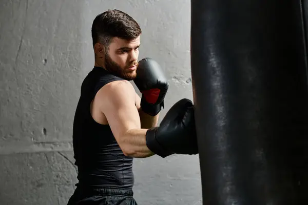 Un hombre guapo con barba, con una camiseta negra y guantes de boxeo, practica sus golpes en una bolsa pesada en un gimnasio. - foto de stock