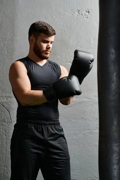 Un hombre guapo con barba se para junto a un saco de boxeo en un gimnasio, practicando técnicas de boxeo con enfoque y determinación. - foto de stock