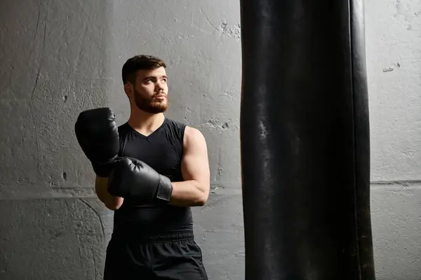 Un hombre guapo con barba se para junto a un saco de boxeo en un gimnasio, practicando boxeo. - foto de stock
