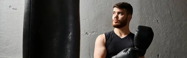 Hombre guapo con barba vistiendo camiseta negra y guantes de boxeo, golpeando ferozmente una bolsa en un gimnasio. - foto de stock