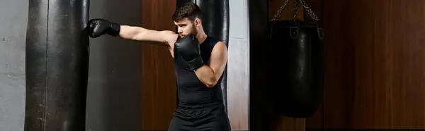 Un bel homme barbu portant une chemise noire et des gants noirs, boxant dans un ring avec un sac de boxe dans un gymnase. — Photo de stock