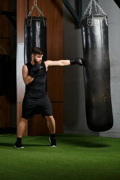 Hombre barbudo guapo con camisa negra y pantalones cortos golpea vigorosamente un saco de boxeo en un entorno de gimnasio. - foto de stock