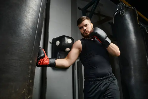Красивый мужчина с бородой в черной рубашке и красных боксерских перчатках бьет боксерскую грушу в спортзале. — стоковое фото