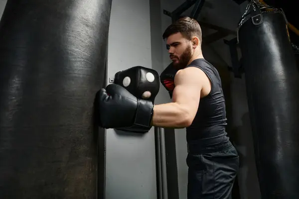 Un hombre guapo con barba, con una camiseta negra y guantes de boxeo, golpea una bolsa en el gimnasio. - foto de stock