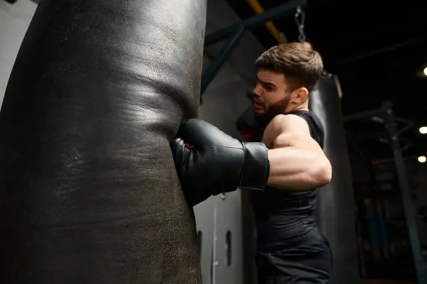 A man with a beard wearing a black shirt and boxing gloves, fiercely punching a bag in a gym. — Stock Photo