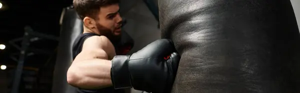 Un hombre guapo con una barba con una camiseta negra y guantes de boxeo, golpeando ferozmente una bolsa en un gimnasio. - foto de stock