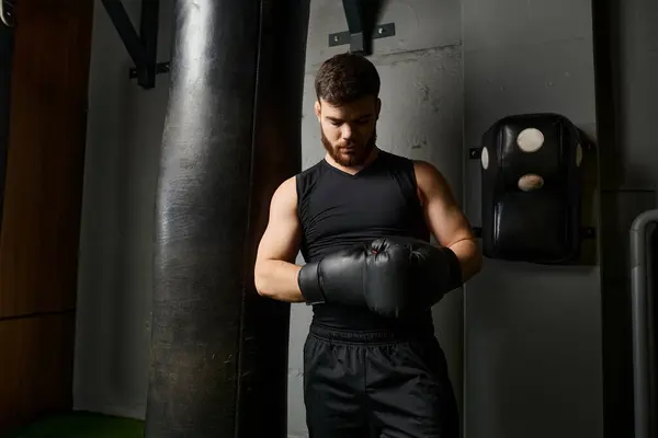 Hombre guapo con barba, con una camiseta negra y guantes de boxeo, golpea ferozmente una bolsa en un gimnasio. - foto de stock