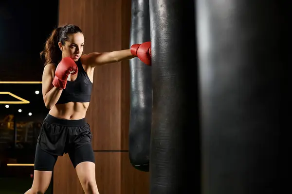 A brunette sportswoman, dressed in a black sports bra, is seen in motion, throwing punches with red boxing gloves in a gym setting. — Stock Photo
