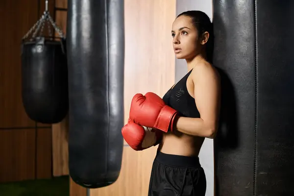 A confident brunette sportswoman in active wear stands next to a punching bag wearing red boxing gloves, ready for a intense workout. — Stock Photo