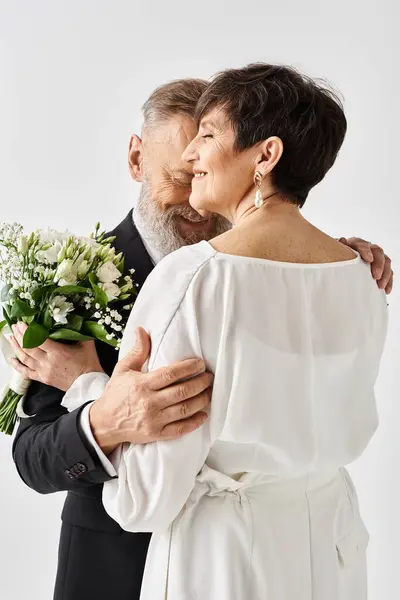 Mariée d'âge moyen et marié en tenue de mariage serrant étroitement tout en tenant des fleurs, célébrant leur journée spéciale dans un cadre de studio. — Photo de stock
